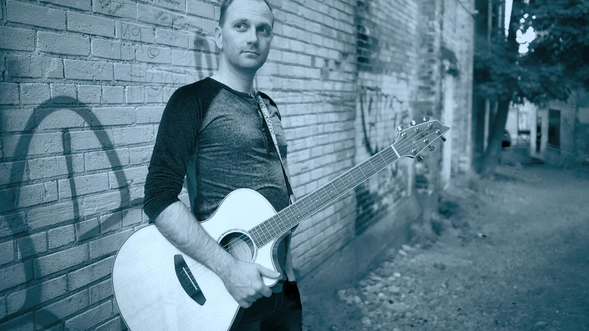 A duotone photo with a man holding a guitar beside a brick building