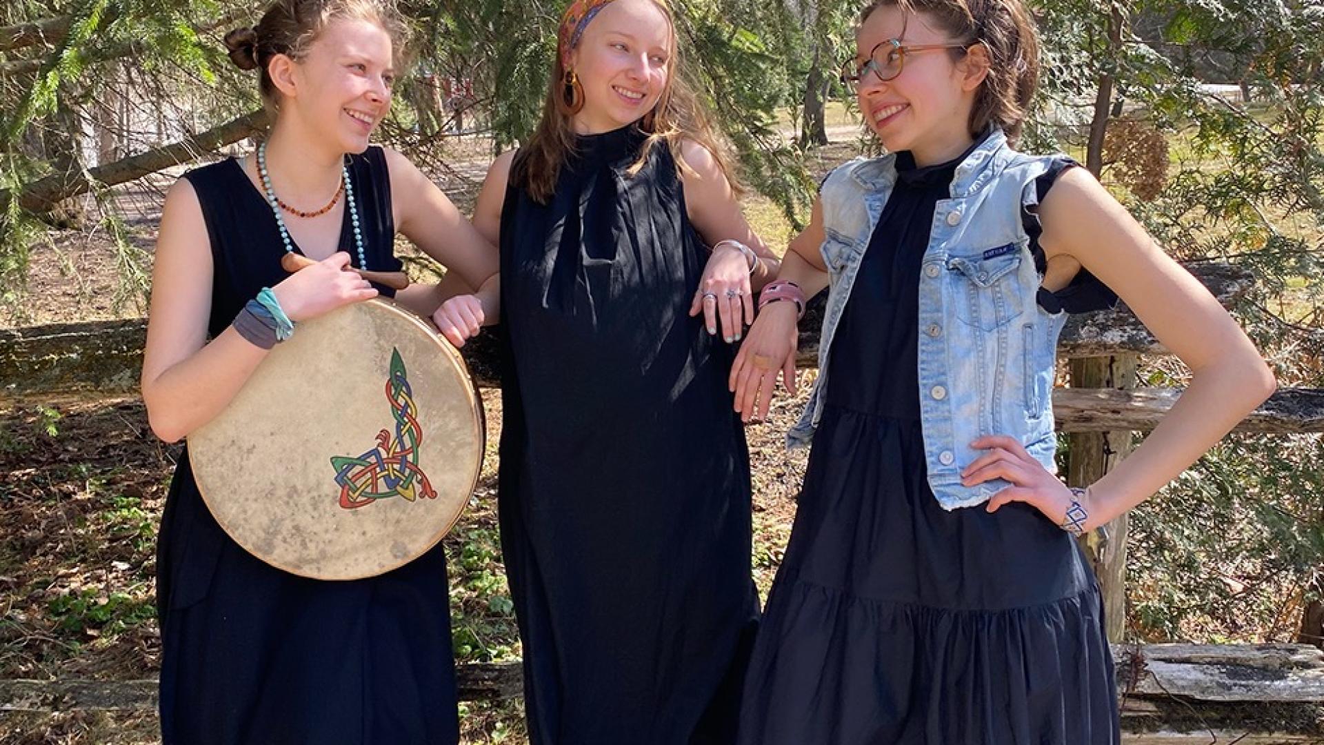 An image of three women in black outfits leaning against a wooden fence smiling under trees