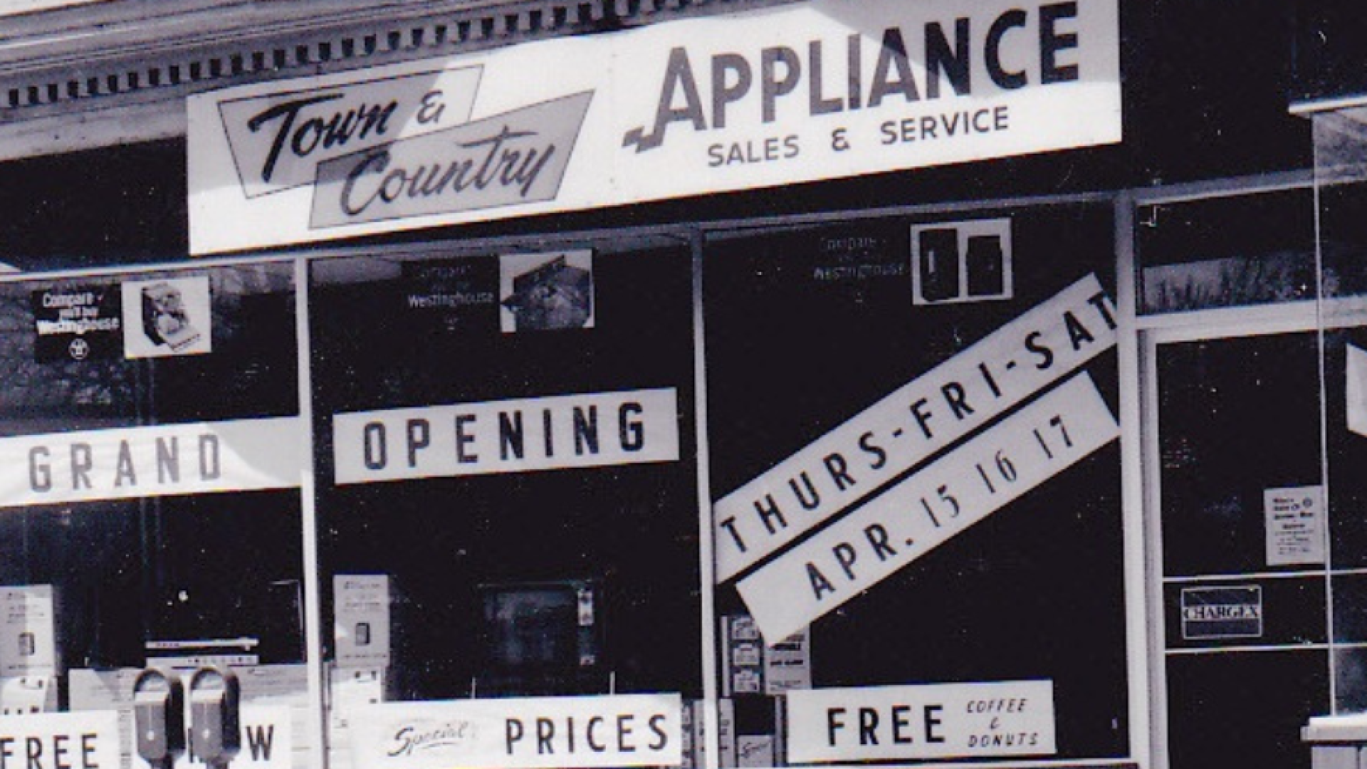 Black-and-white historical photo of storefront windows advertising a grand opening, with large signs reading “APPLIANCE Sales & Service,” “Grand Opening,” and “Thurs–Fri–Sat Apr. 15, 16, 17,” plus a smaller sign for “Free coffee & donuts.” A semi-transparent overlay in the lower left reads “Windows in Time – June 29 to July 31,” and a Hanover, Ontario logo appears in the top left.