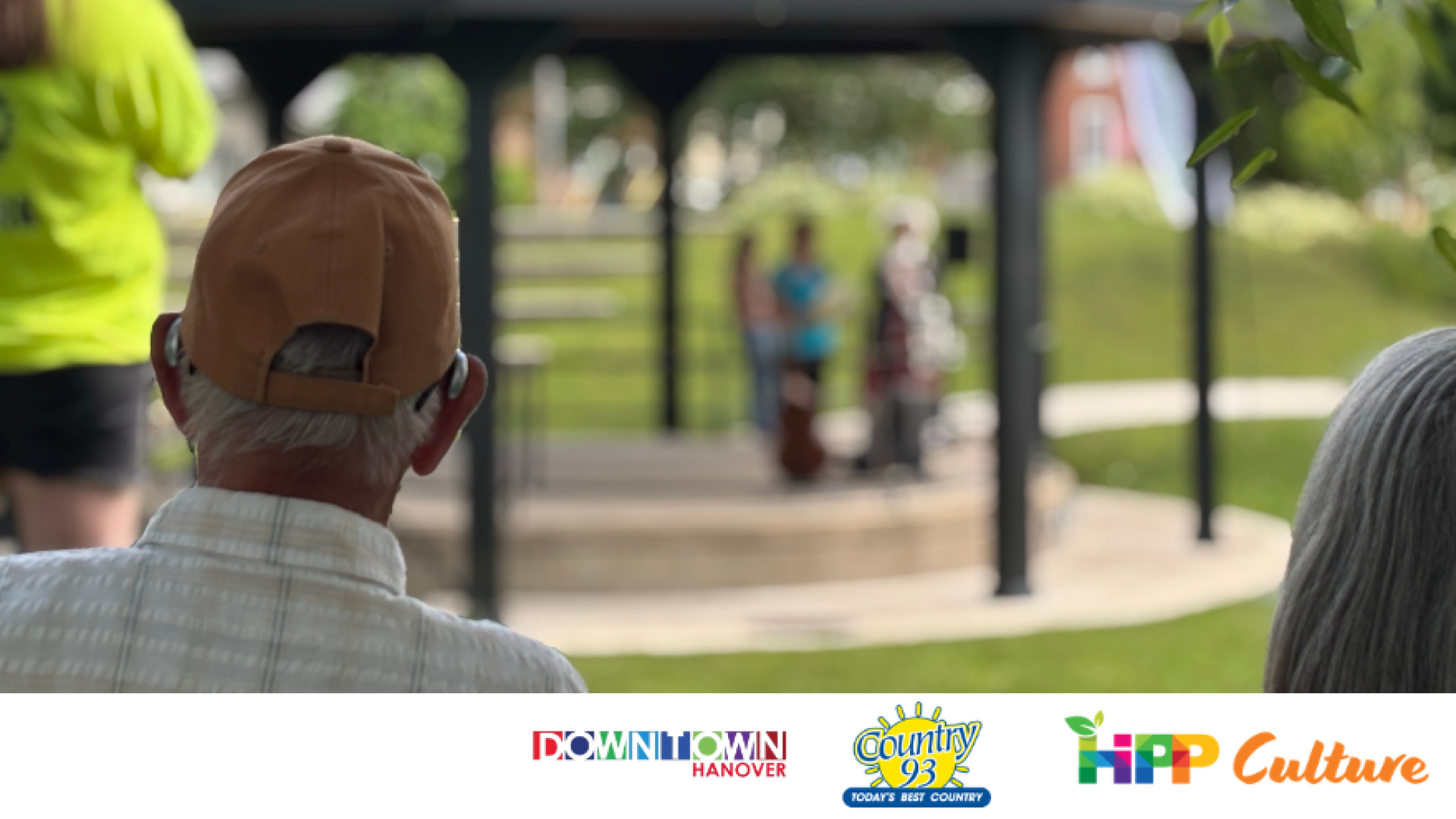 The back of a couple's head, one wearing a baseball cap, watching the bandshell at Heritage Square in Hanover with Downtown Hanover, Country 93 and HIPP Culture Logos