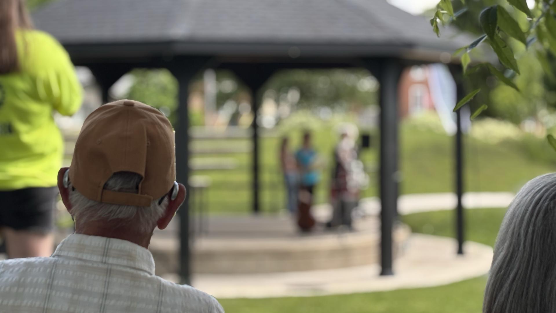 Couple watching bandshell in Heritage Square Hanover, man on the left with a brown baseball cap, grey hairs and a white and brown plaid shirt, the woman is sitting in a red chair and has grey hair, the photo is of the backs of their heads, in front of them is green space and a black roofed bandshell
