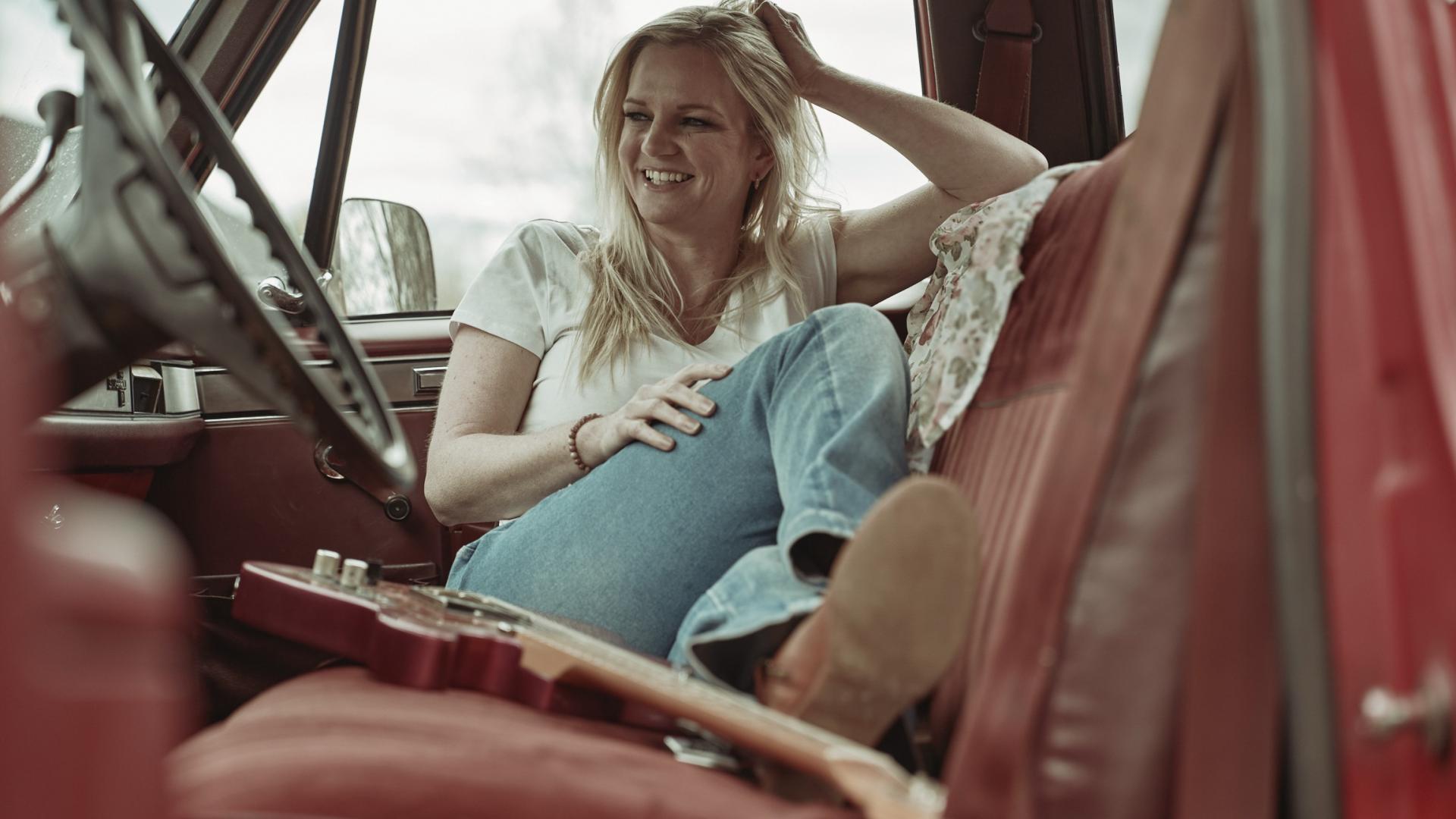 Photo of Lisa McEwen, a female with blonde hair, wearing a white shirt and blue jeans, sitting in the passenger seat of a red car smiling beside her acoustic guitar