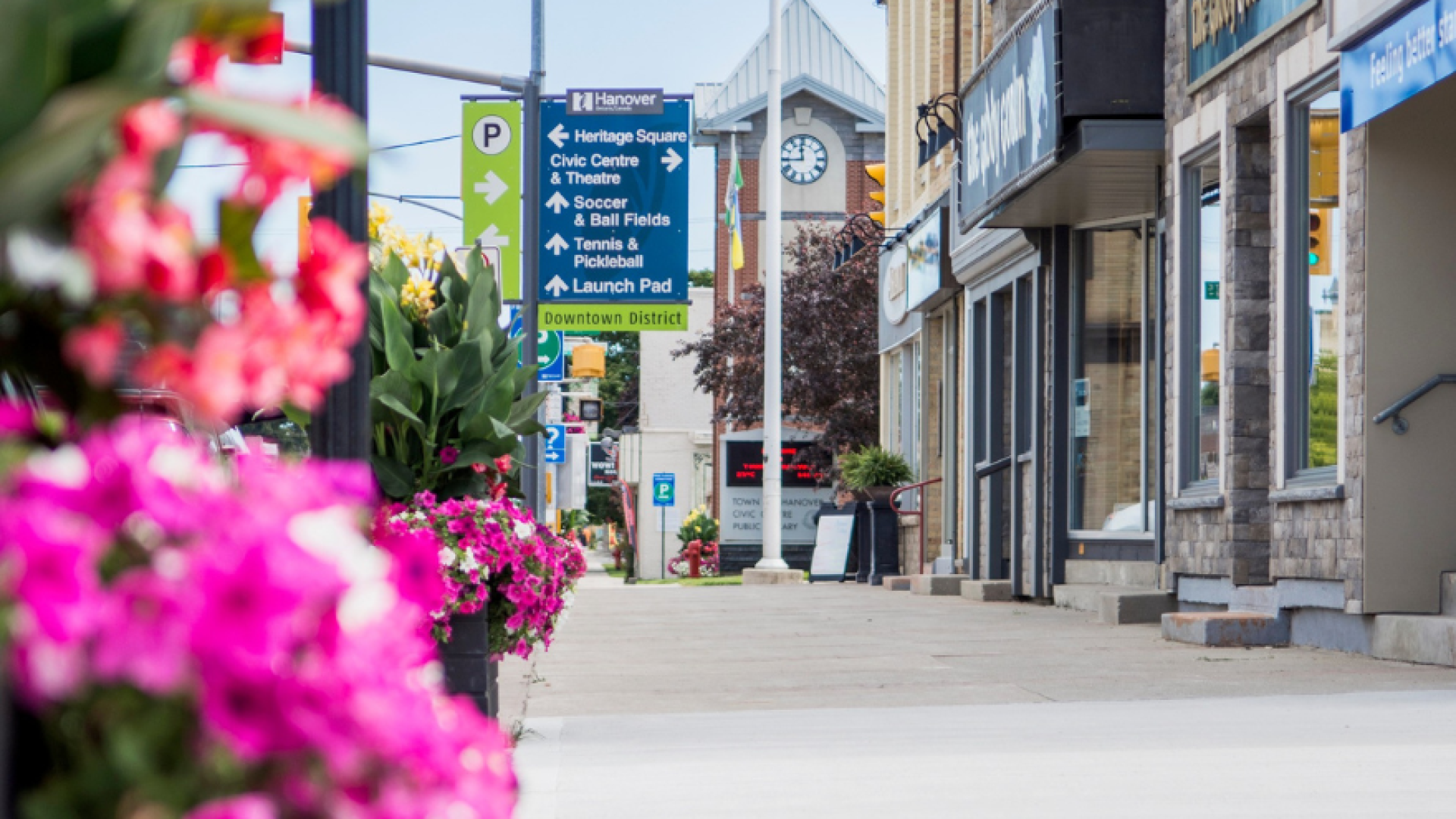 A photo of downtown Hanover with flowers in the foreground, directional signage in the middle ground, alongside business storefronts, with Hanover Civic Centre in the background.