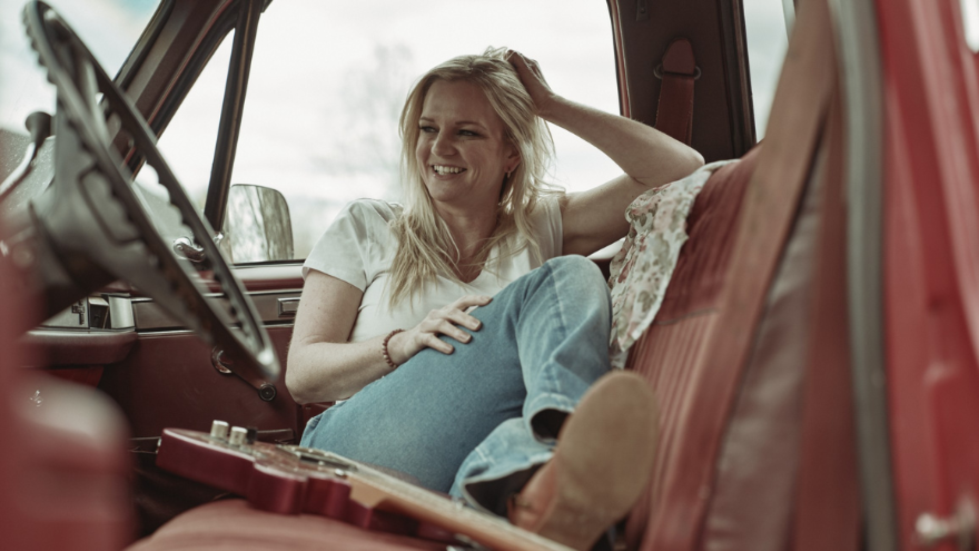 a photo of Lisa McEwen, blonde hair, in a white shirt and blue jeans, sitting in the passenger seat of a car with red leather seats, beside an acoustic guitar
