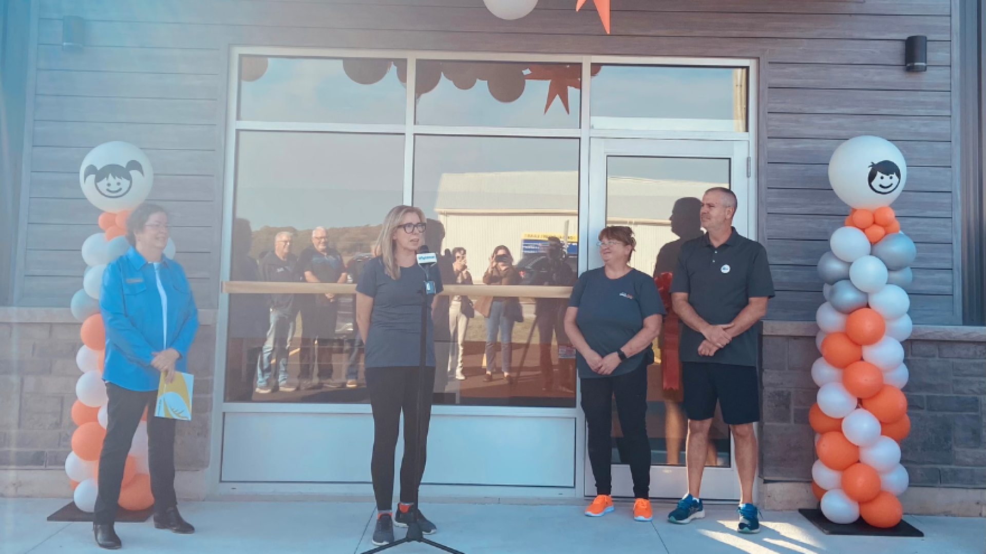A group of people standing in front of a building made of wood and brick, with orange, white and silver vertical balloon stands, a woman in a blue jacket is smiling with her arms folded over each other, a blonde woman with black glasses is speaking into a micorophne and two additional worked are standing in the background with their hands crossed. It is a celebration.