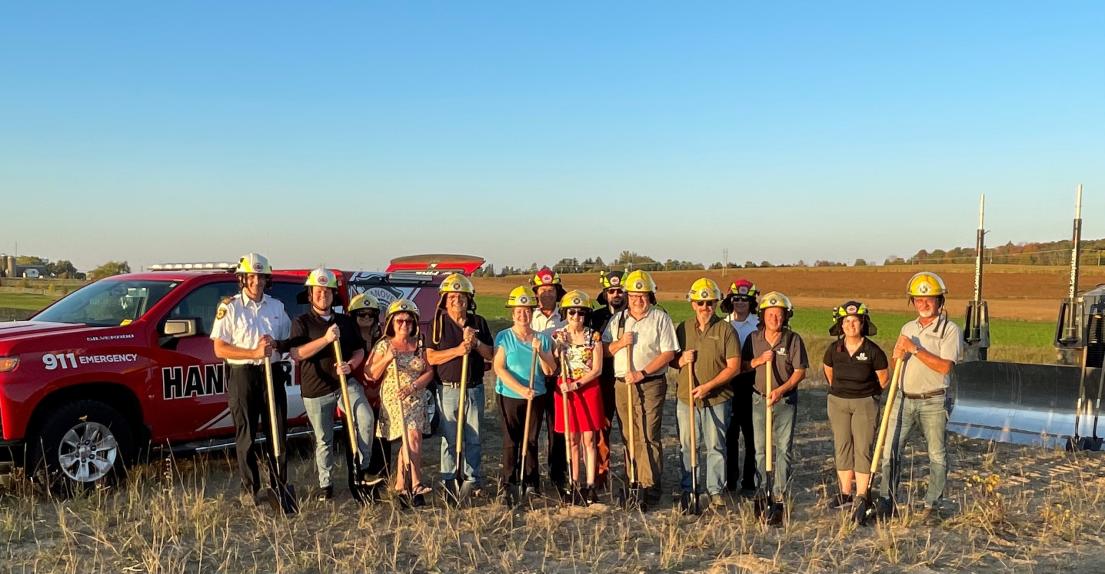 Group of people holding shovels. They are standing in a empty field in front of a fire emergency vehicle.