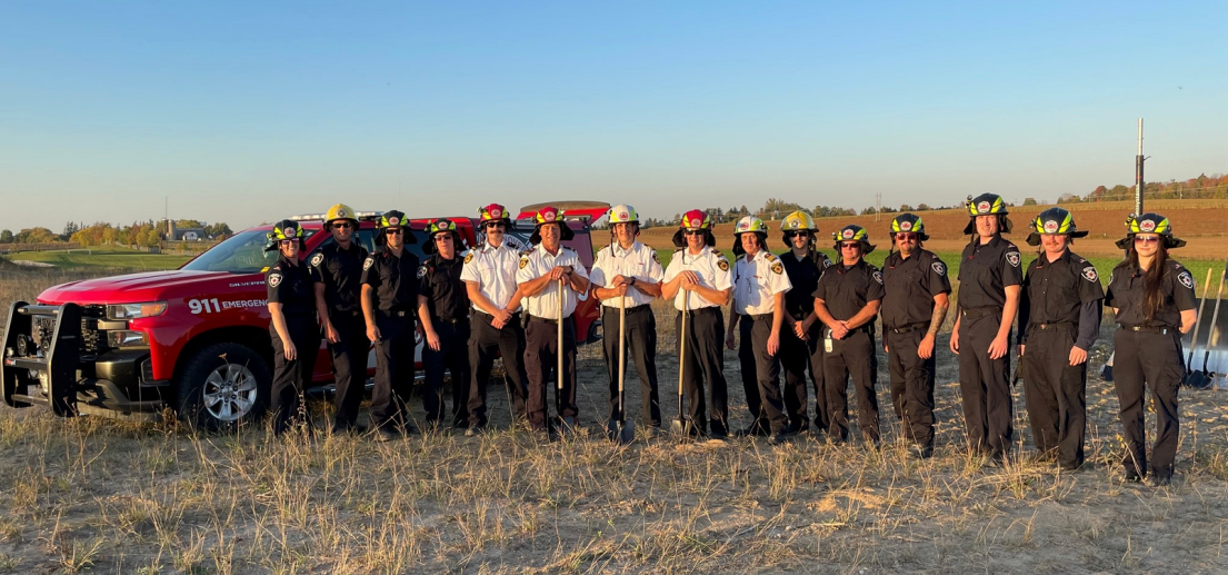 Group of people holding shovels. They are standing in a empty field in front of a fire emergency vehicle.