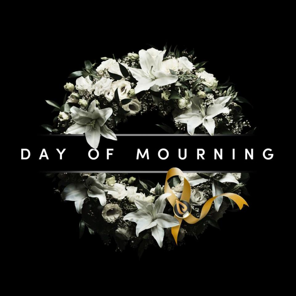 White lilies and roses arranged in a circular wreath with a yellow ribbon on a black background, captioned 'Day of Mourning'.
