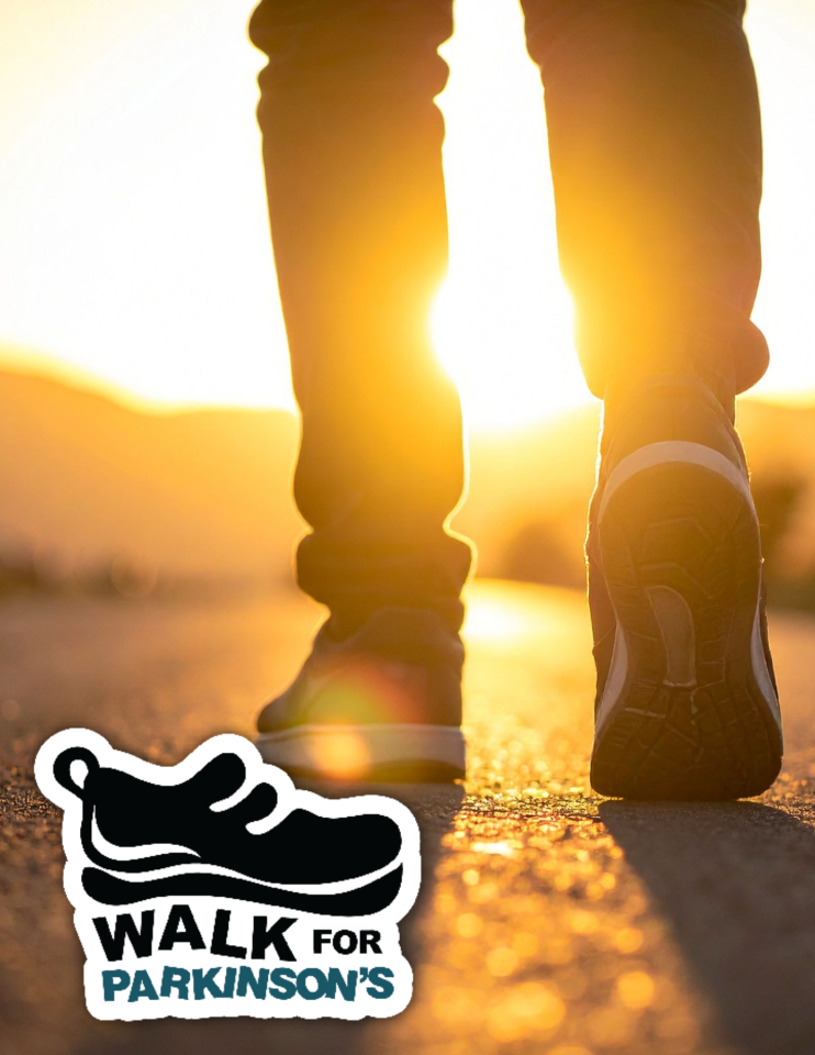 Someone walking on the pavement towards a rising sun, wearing running shoes and jeans, close up from knees down and a walk for parkinson's logo
