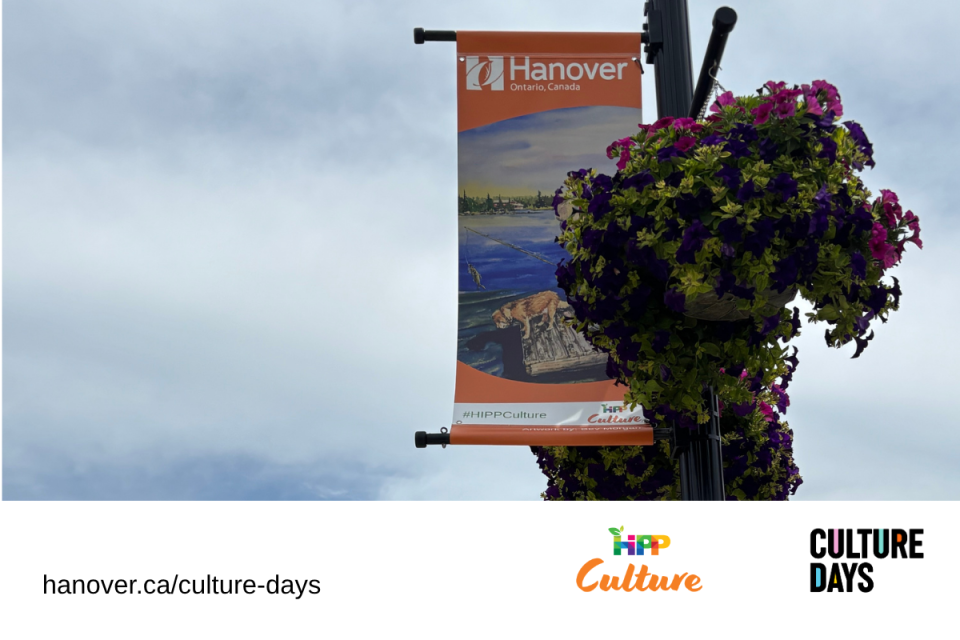 A photo of a cloudy day, with an art banner that has the Town of Hanover logo and is hanging on a post beside a flower basket with purple flowers.