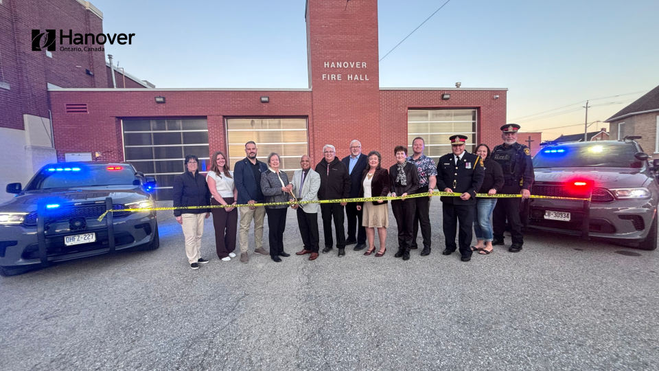 A group of 13 people standing in front of Hanover Fire Hall behind caution tape with two police cruisers.