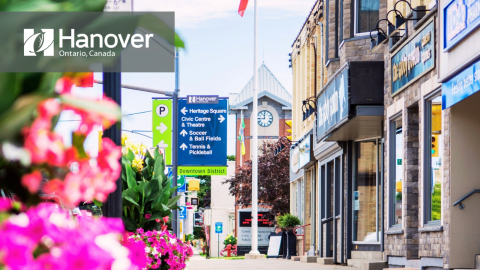 Downtown Hanover with pink and purple blooming planters on the sidewalk, images of store front, directional signage in the background along with Canadian Flag and the Hanover Clock Tower and Town of Hanover Logo