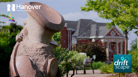 Heritage Square fountain with water spouting out with the Hanover Civic Centre in the background and the Pride Flag with white Town of Hanover Logo in the top left corner and Driftscape logo in the bottom right corner