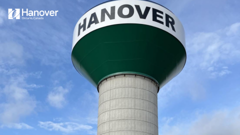 Composite Water tower with dark green underside and a white front, with the text, Hanover on it and a clear blue sky with light cloud coverage, also has a white Town of Hanover Logo