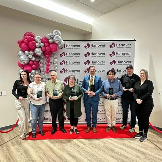 A group of 8 people holding awards in front of a pop up banner that says Hanover Chamber of Commerce. There is a red carpet on the ground and pink and silver balloons.